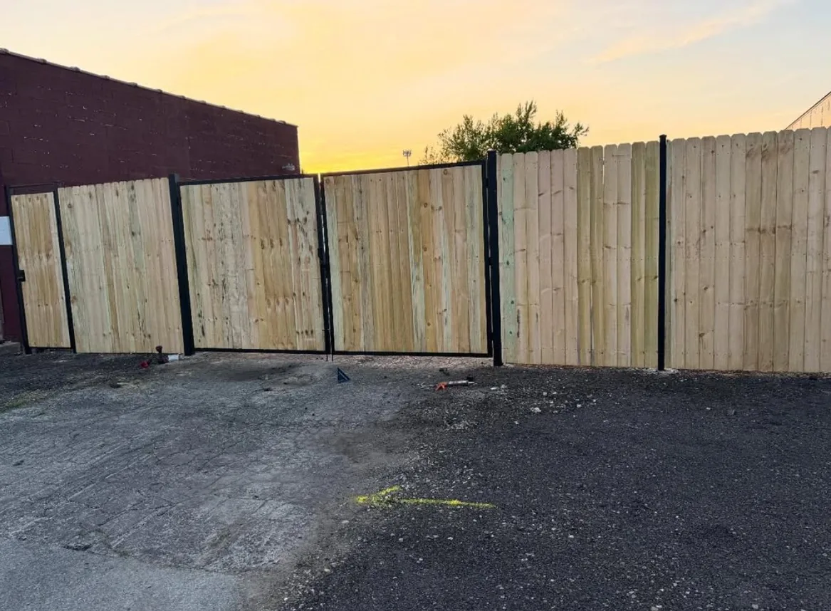 Double drive gate framed in black steel with wood privacy panels, closing off a commercial lot at sunset next to a matching wood stockade fence