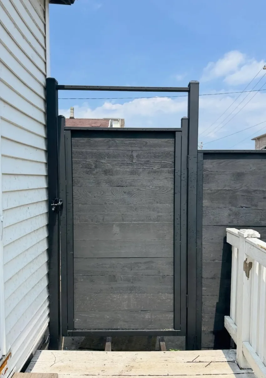 Custom walk gate built from dark stained horizontal wood planks in a welded black steel frame, hung with a latch between a house and a matching privacy fence