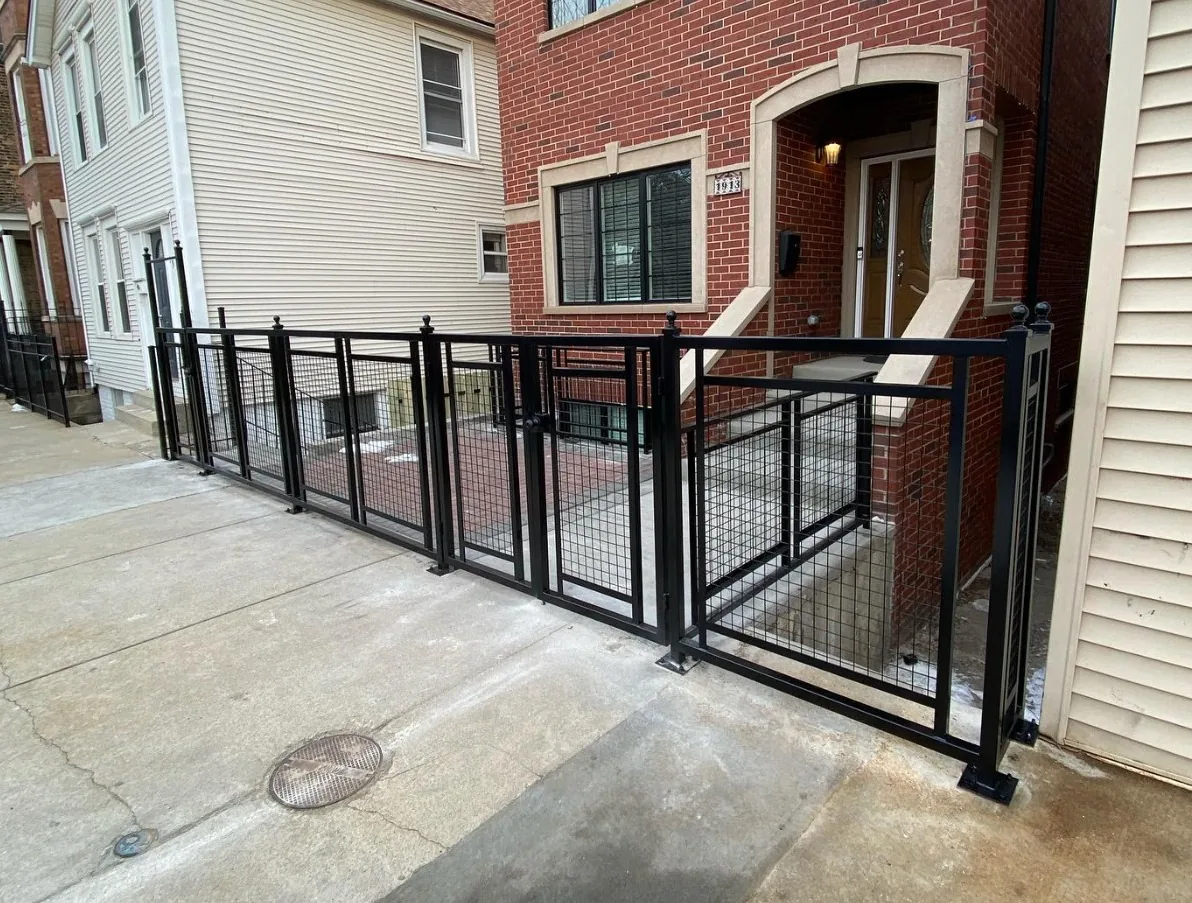 Black steel fence with welded wire mesh panels and a matching walk gate, installed along the concrete gangway of a red brick two-flat