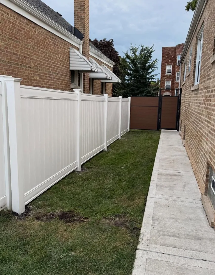 White vinyl privacy fence panels running down a grass side yard between two brick buildings, with a brown composite gate at the far end